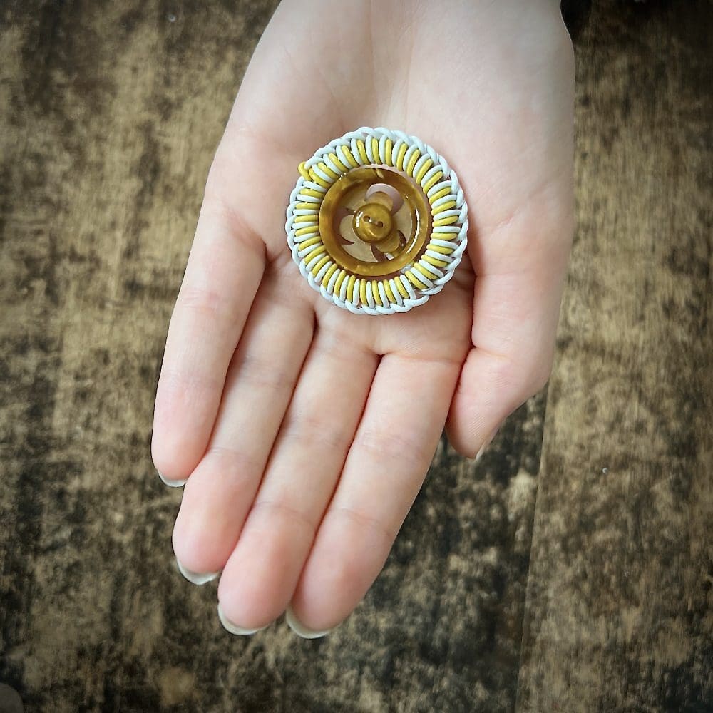 A circle brooch of yellow and white wire twisted in a decorative fashion is shown in the palm of a hand. The centre of the brooch is an amber vintage button.
