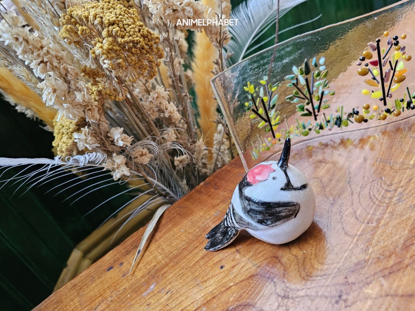 Ceramic lesser spotted woodpecker sat on a wooden table, with dried flowers behind and glass art to the side