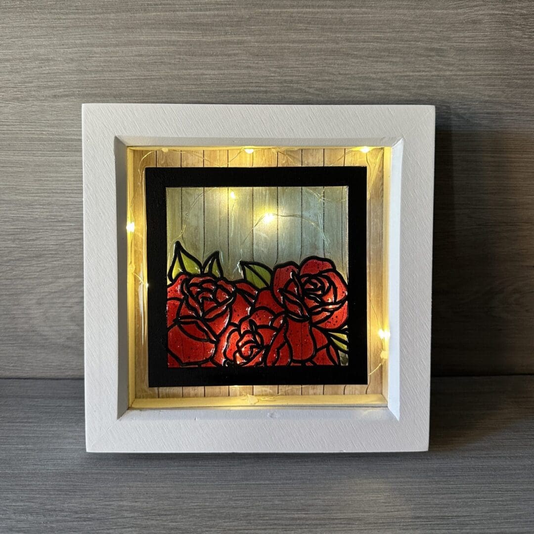 a 6 inch white box frame with string of lights inside. On the front is a black square frame with roses detail. The roses are infilled with red resin