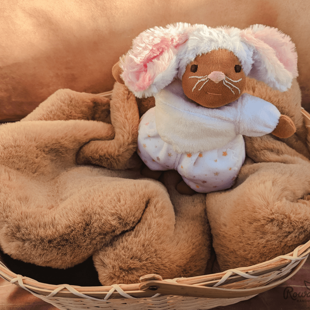 little mouse doll with brown skin and a pink embroidered nose lying in a wicker basket on a brown furry blanket, with a cinnamon background