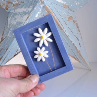 Dark blue wooden frame with two clay daisies on wire inside
