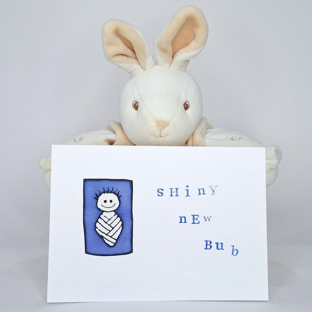 Cute baby bunny holds up greetings card with a cartoon baby in a papoose, the card reads 'Shiny New Baby'