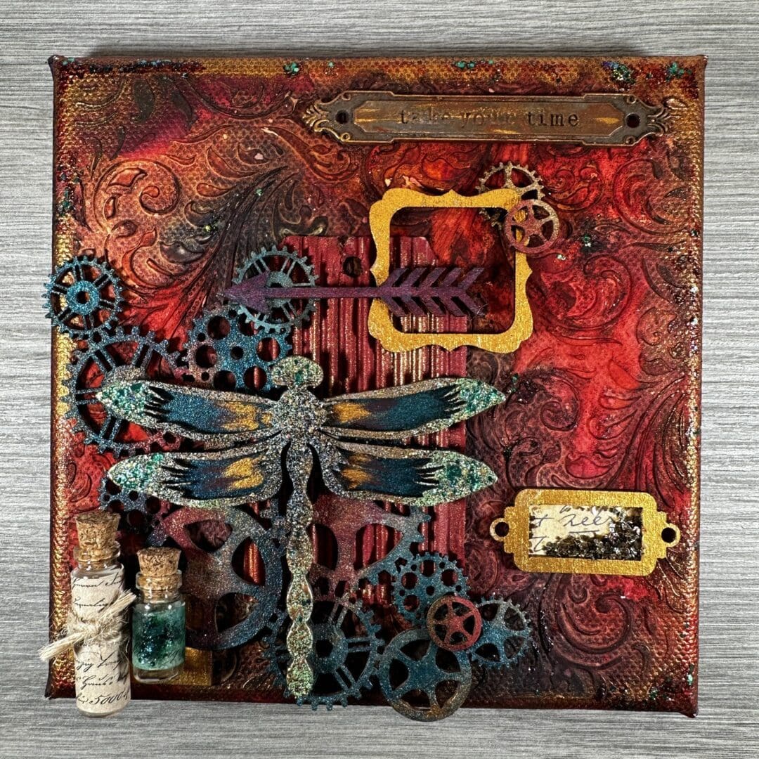 6 inch mixed media canvas in rust tones with a textured background. Layered up with small wooden cogs and embellishments topped with a dragonfly in tones of turquoise.