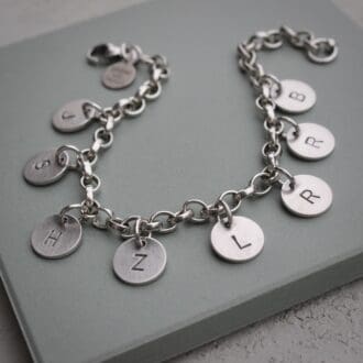 A sterling silver chain charm bracelet with a series of round disc tags each stamped with a single initial, on a green tile