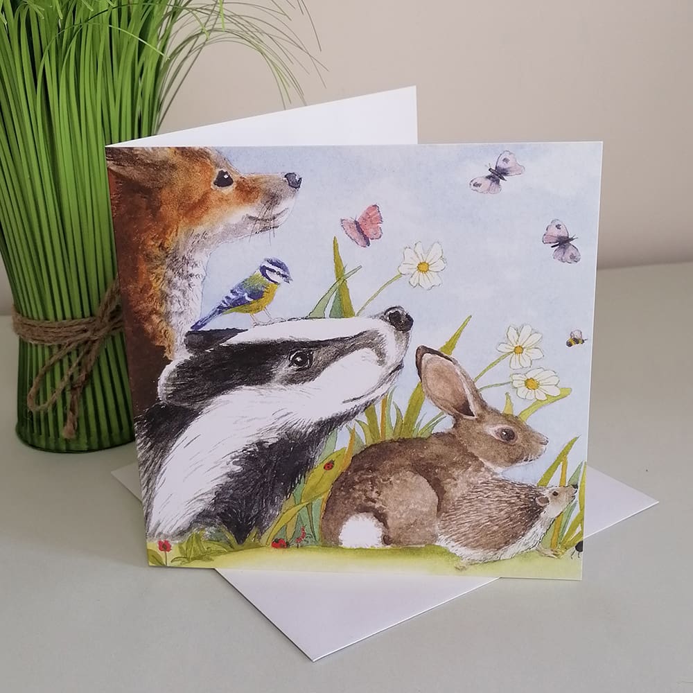 Square greetings card featuring a group of British wild animals and insects including a brown fox, rabbit, hedgehog blue tit bird and badger, sat together amongst daisies and grassland.