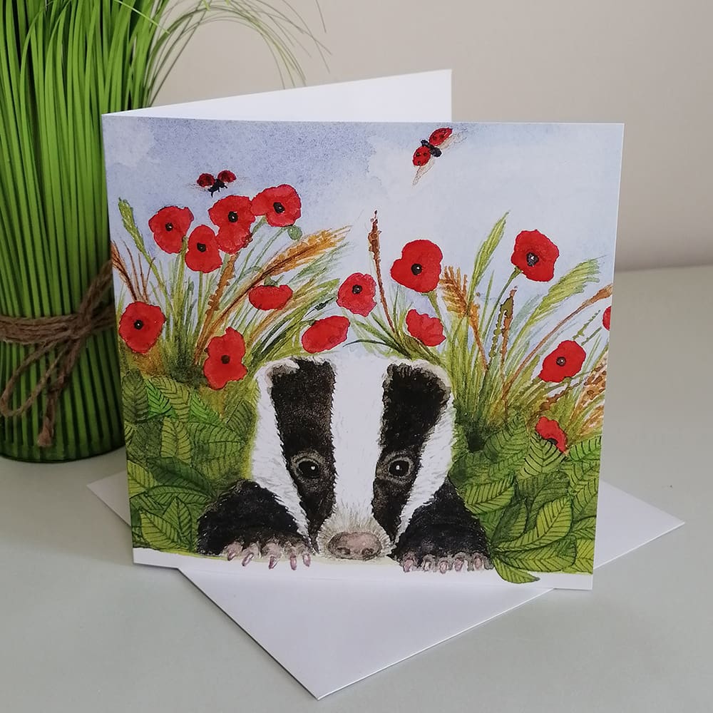 Square greetings card featuring a young badger cub resting amongst the leaves and red poppies. The backdrop of the scene is blue sky and flying ladybugs.