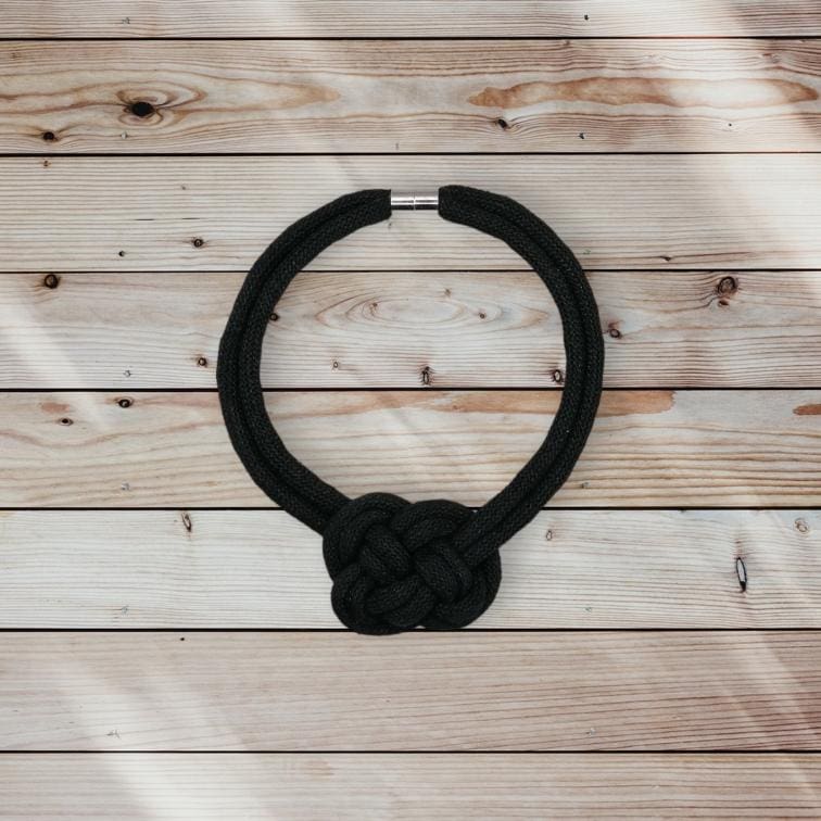 Flatlay view of black rope necklace with central knot feature which is laying on a light coloured pine background