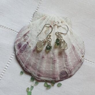 a pair of dainty drop earrings made from small pieces of sea-glass wrapped in fine silver wire, in white and green, hung from a silver shepherds hook - placed on top of a large pink and white bivalve shell surrounded by tiny pieces of sea glass