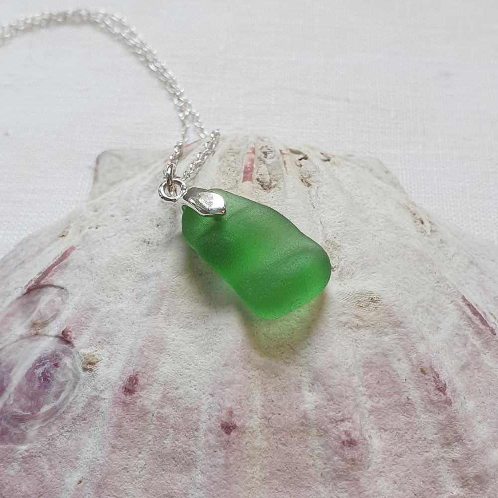 An emerald green bumpy piece of sea glass with a silver bail and chain necklace sitting on a large shell for display.