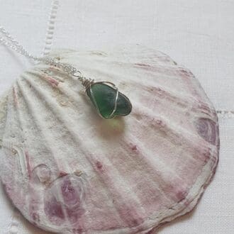 a blue-green fine silver wire wrapped piece sea glass necklace on a sterling silver chain displayed on a large pink and cream bivavle shell.