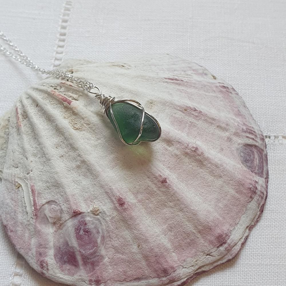 a blue-green fine silver wire wrapped piece sea glass necklace on a sterling silver chain displayed on a large pink and cream bivavle shell.