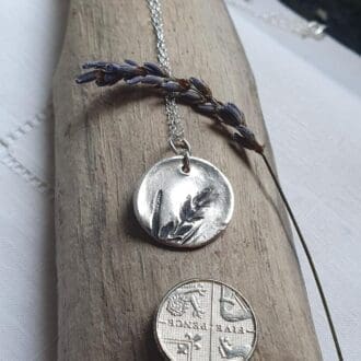 A fine silver disc imprinted with a lavender stem, resting on a piece of driftwood with a 5 pence piece alongside for scale.
