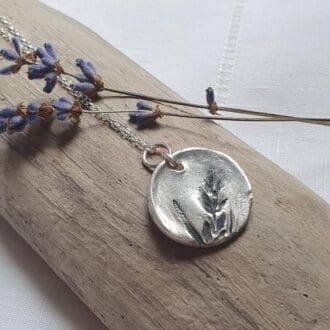 A solid silver disc pendant imprinted with lavender leaves and flowers which have been aged. sitting on a piece of driftwood with lavender flowers