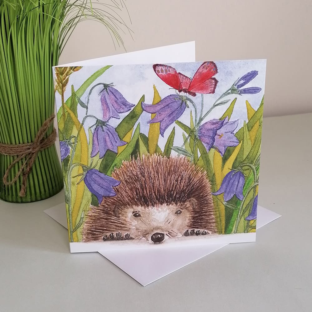 Square greetings card featuring a young hedgehog with a bright pink butterfly amongst blue harebell flowers.