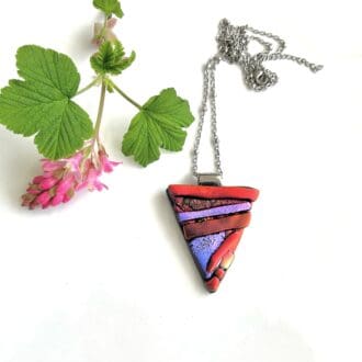 A triangle shaped fused glass pendant in bright red and purple metallic look glass on a white background with a pink flower placed beside it for decoration. A stainless steel satellite chain.