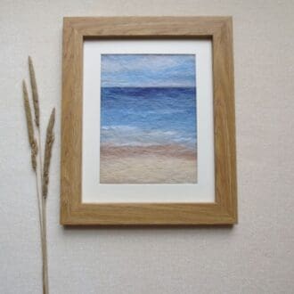 A needle felted wool painting of a sand and sea coastal seascape in a light oak effect frame.