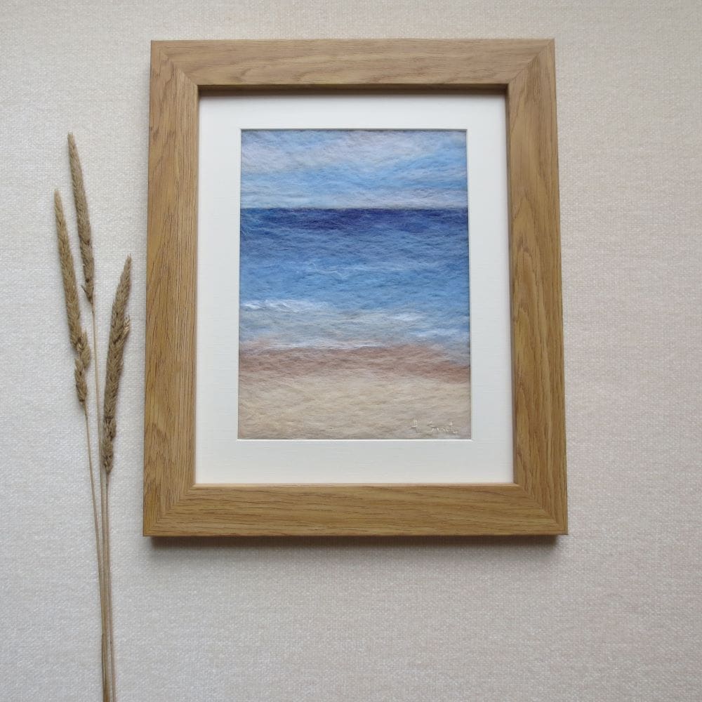 A needle felted wool painting of a sand and sea coastal seascape in a light oak effect frame.