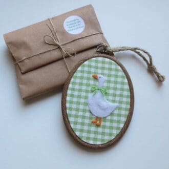Needle felted white goose on an apple green gingham fabric and in a wood effect presentation hoop with jute hanging cord.