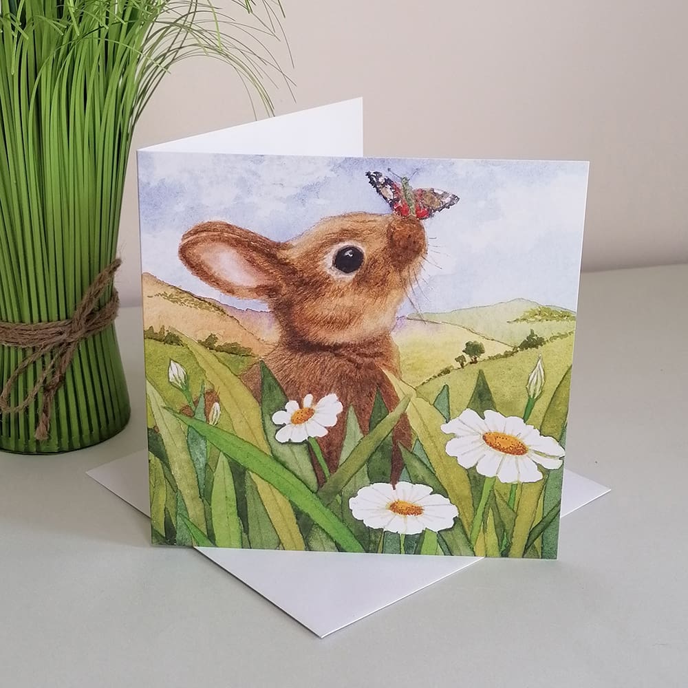 Square greetings card featuring a young brown rabbit with a butterfly landing on his nose sat amongst daisy flowers. The background of the card is set in the countryside.