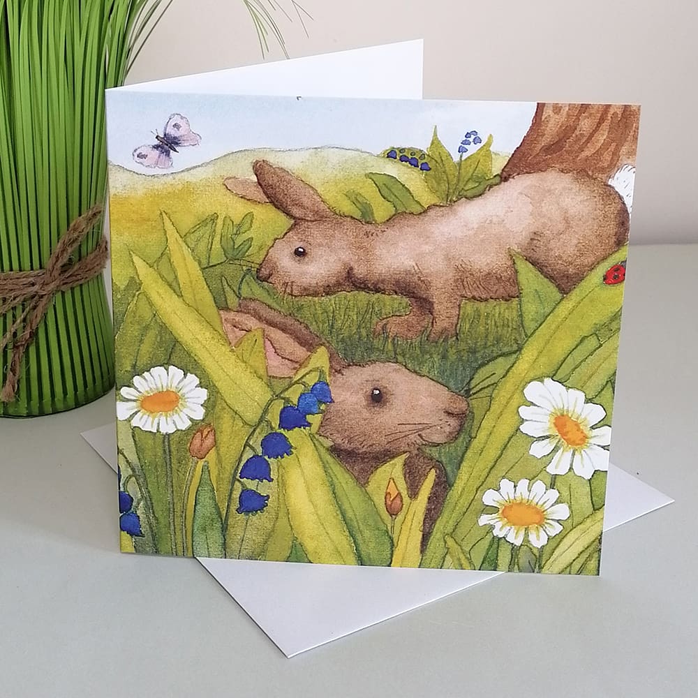 Square greetings card featuring two brown rabbits nibbling grass The background of the card is set in the countryside.