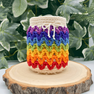 a small jar covered in rainbow crochet stripes. It is sittiing on a wood slice and there is greenery in the background