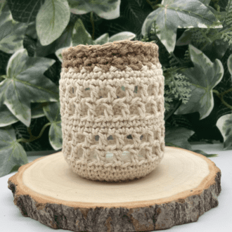 a little glass jar covered in a lacey crochet pattern with jute finish round the neck of the jar. It's standing on a log slice and there is greenery in the background