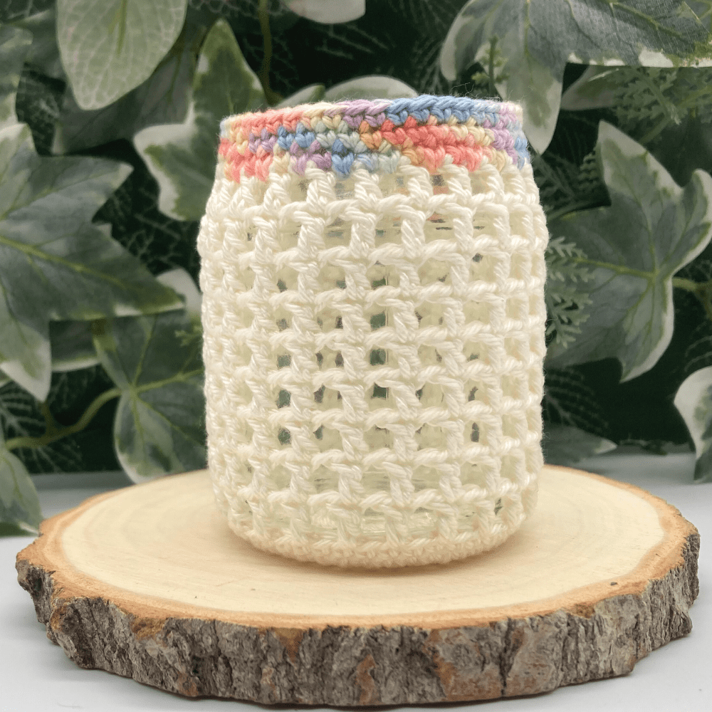 a little glass jar decorated with a cover of cream crochet with a pastel coloured rim. It is standing on a wood slice and there is greenery behind