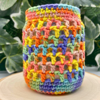 a little glass jar covered in multicolourred lacey crochet. It is standing on a log slice and there is greenery in the background