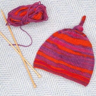 A top knot beanie for 1 or 2 year old child lying alongside a pair of bamboo knitting needles and a ball of wool.