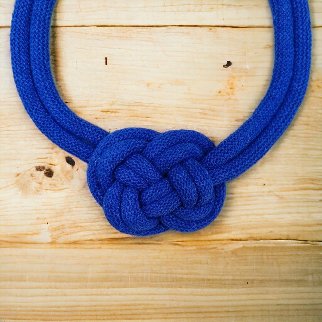 Close up of knot detail in chunky blue statement rope necklace, shown against a light wood background.