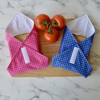 Two sandwich wraps placed on a chopping board with a sandwich wrapped inside. The left wrap is pink with small white dots. The right wrap is blue with small white dots