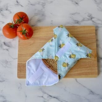 Sandwich wrap on a wooden chopping board, open at one end with a sandwich inside. Fabric is light blue with yellow and black bumble bees