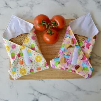 Two sandwich wraps on a wood chopping board, open at one end with a sandwich inside. Left wrap is light yellow with ice lollies and fruit. The right wrap is light pink with cupcakes