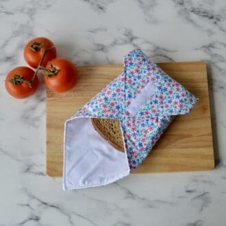 Reusable sandwich wrap resting on a wooden chopping board with a sandwich wrapped inside. Made from cotton fabric with small pink and blue daisies