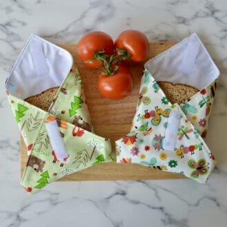 Two sandwich wraps on a wooden chopping board with a sandwich inside. Left sandwich wrap is green with woodland animals. Right wrap is cream with garden bugs
