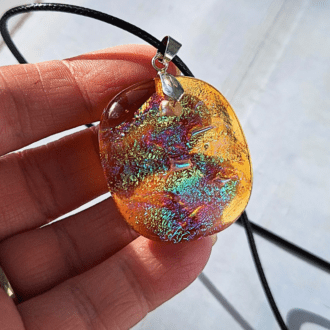 A close-up photo of a handmade orange dichroic glass pendant. The orange glass shimmers with flashes of purple and blue depending on the angle of light. The pendant has a silver-plated pinch bail and hangs from a black corded necklace.