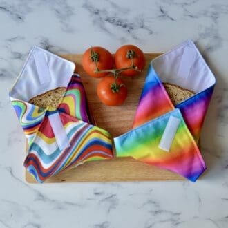 Two Rainbow sandwich wraps laying on a wooden chopping board. The left sandwich wrap in a retro rainbow design and the right is rainbow stripes. Sandwich wraps are folded over a sandwich