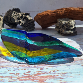 A close-up photo of a fused glass leaf sculpture in shades of green and blue with a textured surface. The leaf has a slightly curved shape and sits on a wooden table.