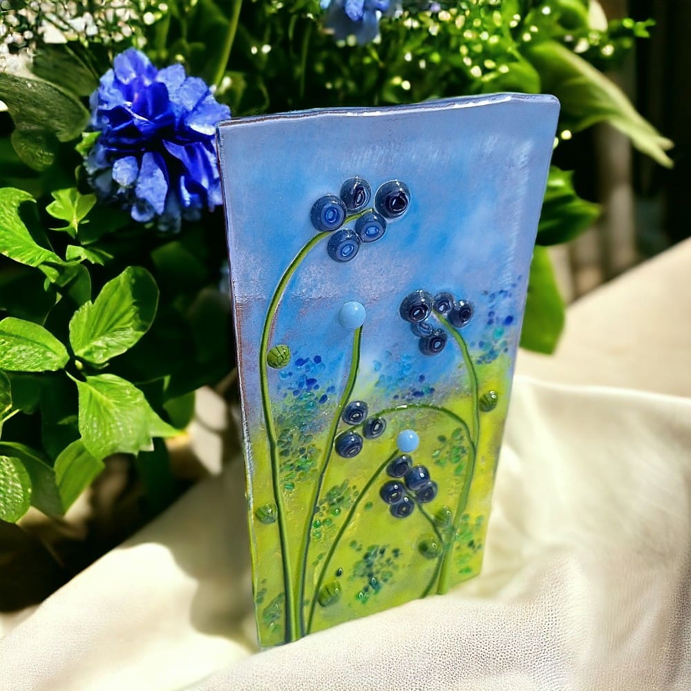 A close-up photo of a blue fused glass tealight holder. The tealight holder has an abstract floral design in various shades of blue. The tealight holder sits on a white background.