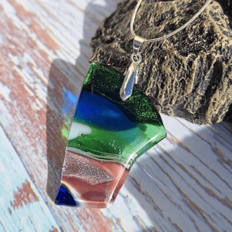 A close-up photo of a handmade rectangular dichroic glass pendant in shades of greenand blue The pendant shimmers with flashes of purple and blue depending on the angle of light. It has a silver-plated pinch bail and hangs from a delicate silver-plated snake chain.