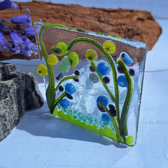 A close-up photo of a square fused glass tealight holder. The design features a vibrant blue meadow scene with green accents, reminiscent of a summer day. The tealight holder sits on a white background.