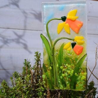 A close-up photo of a handmade fused glass plant stake featuring a cluster of yellow daffodils with green leaves and grass, attached to a stainless steel rod.