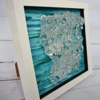 A close-up photo of a textured glass wall art piece titled "Dancing in the Waves." The artwork features a blue and green acrylic background with a clear textured glass overlay, resembling waves in motion. The piece is framed in a white frame.