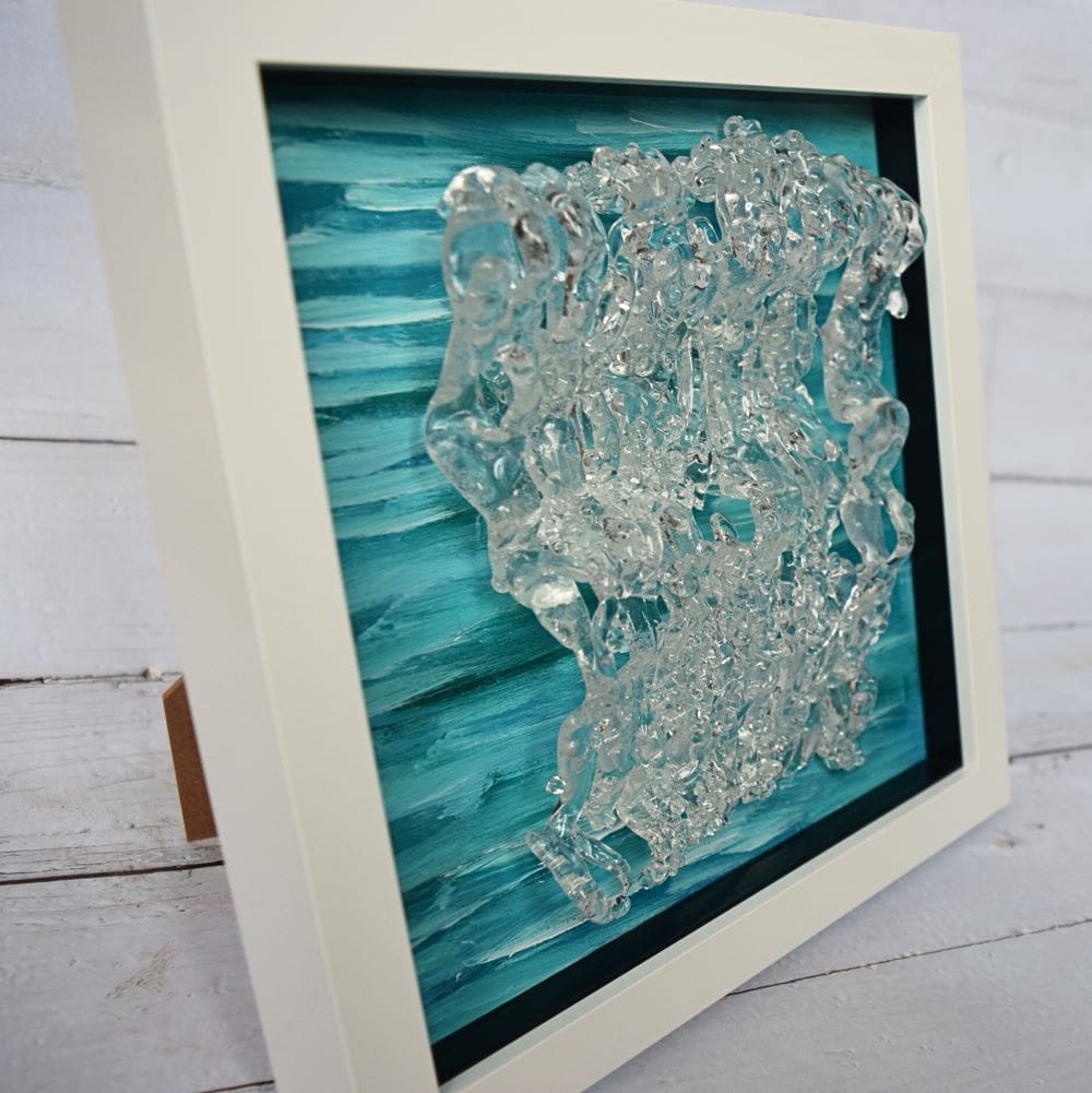 A close-up photo of a textured glass wall art piece titled "Dancing in the Waves." The artwork features a blue and green acrylic background with a clear textured glass overlay, resembling waves in motion. The piece is framed in a white frame.