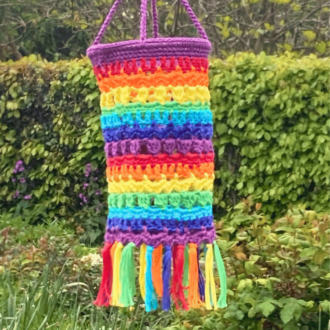 a crochet lantern in rainbow stripes. The bottom is finished with a fringe also in rainbow colours. It is hanging in the garden in front of a flower bed of green and spring colours