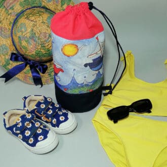 Kids red top duffle bag with sail boat print and navy blue base ready for the beach. Yellow kids swimsuit, blue flowery shoes and a summer straw hat decorated with a blue ribbon