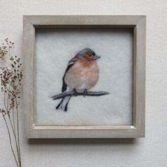 A handmade needle felted wool picture of a peach coloured chaffinch on a cream wool background and in a pale natural wood box frame.