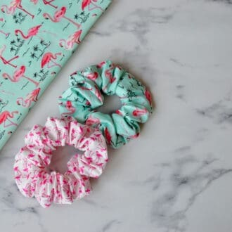 Two scrunchies are shown. One is light green and adorned with pink flamingos, while the other is white, also featuring pink flamingos.