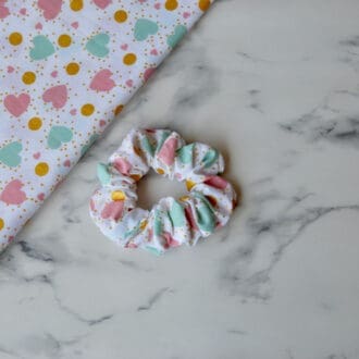 A white cotton scrunchie decorated with green and pink hearts, interspersed with gold spots.