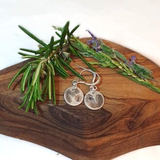 pair of silver hawthorn imprint earrings displayed with herbs on a wooden plinth